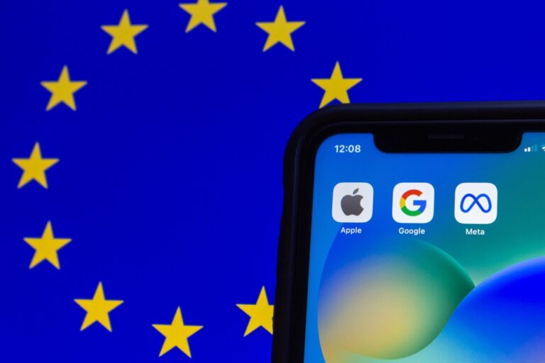 Die Flagge der EU erstreckt sich im Hintergrund. Sie wird in der rechten unteren Bildecke von einem Smartphone überdeckt, auf dessen Bildschirm die App-Symbole von „Apple“, „Google“ und „Meta“ zu sehen sind.